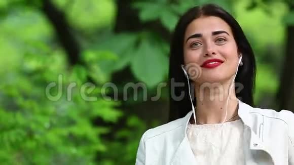 一位女士在公园里听着一部带耳机的智能手机里的音乐一位美丽的女士用带耳机的智能手机听音乐视频的预览图