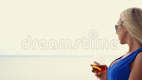 美丽的金发碧眼的金发碧眼正在喝着酒杯里的白葡萄酒欣赏着海洋的地平线站在阳台上视频的预览图