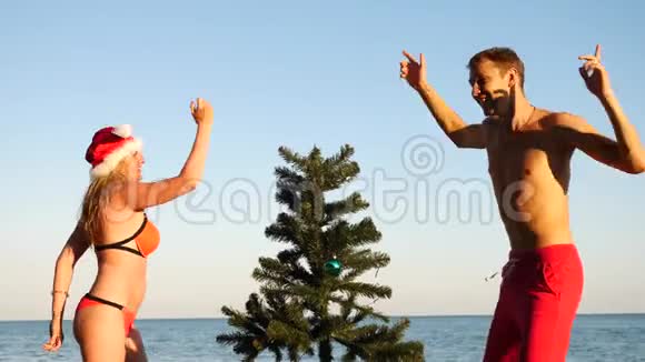 两个戴着圣诞帽的男人和女孩在海滩上迎接新年和圣诞节把圣诞树装饰在圣诞树上视频的预览图
