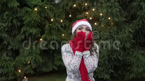 戴着红领巾和手套的圣诞老人帽子的快乐的女人在圣诞树的背景下跳跃视频的预览图