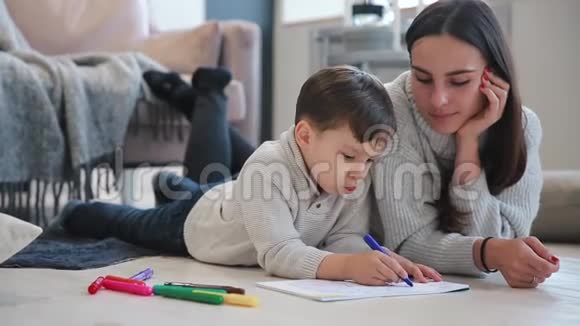 美丽的年轻母亲穿着一件暖和的毛衣躺在地板上我的儿子在纸上画着记号描绘着他的形象视频的预览图