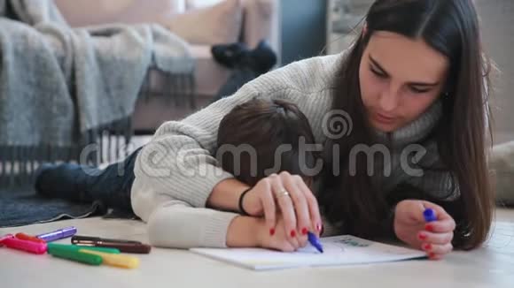 美丽的年轻母亲穿着一件暖和的毛衣躺在地板上我的儿子在纸上画着记号描绘着他的形象视频的预览图