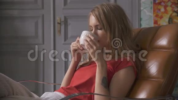 一个年轻的女人端着茶坐在家里的扶手椅上视频的预览图