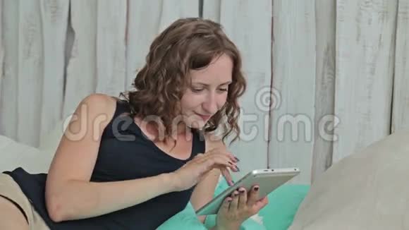 一位年轻的卷发女士躺在床上拿着平板电脑视频的预览图