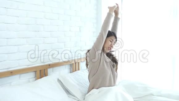 快乐美丽的年轻亚洲女人早上醒来坐在床上伸展在舒适的卧室透过窗户视频的预览图