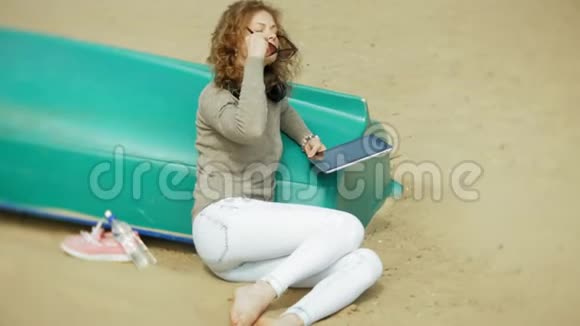 年轻漂亮的女人坐在海滩上戴着耳机坐在船附近用平板电脑视频的预览图