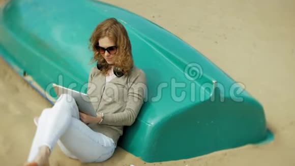 年轻漂亮的女人坐在海滩上戴着耳机坐在船附近用平板电脑视频的预览图
