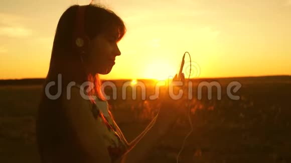 快乐的女孩听音乐在美丽的夕阳的光芒中跳舞戴着耳机的年轻女孩视频的预览图