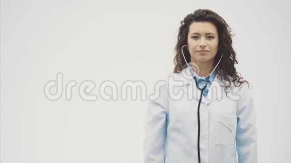 幸福的年轻微笑女孩医生的肖像穿着白色长袍站在灰色的背景上在此期间视频的预览图