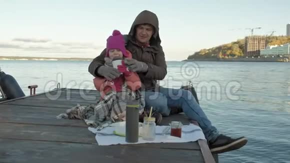 男人和女孩父亲和女儿坐在河港喝茶野餐笑穿着暖和的衣服视频的预览图