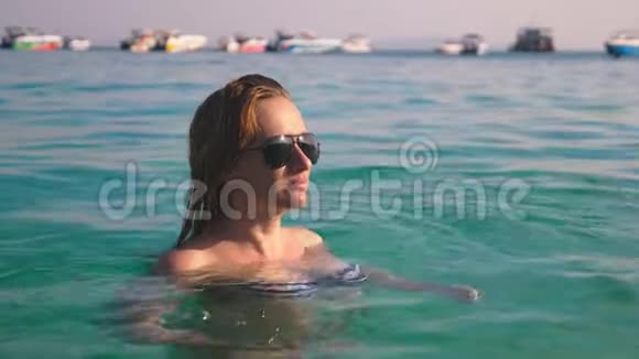 穿着泳衣和黑色太阳镜的金发女孩在蓝色海水中游泳的美丽模特视频的预览图