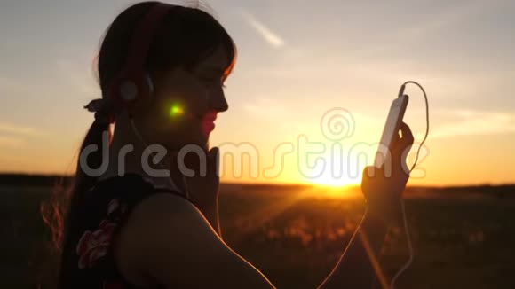 快乐的女孩在美丽的夕阳下在智能手机上听音乐戴着耳机的年轻女孩视频的预览图