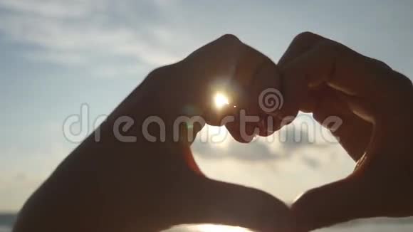女孩用她的双手在海洋背景上用美丽的金色夕阳做心女性手臂的剪影视频的预览图
