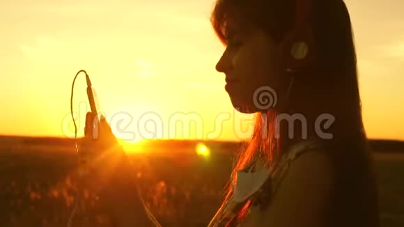 快乐的女孩听音乐在美丽的夕阳的光芒中跳舞戴着耳机的年轻女孩视频的预览图