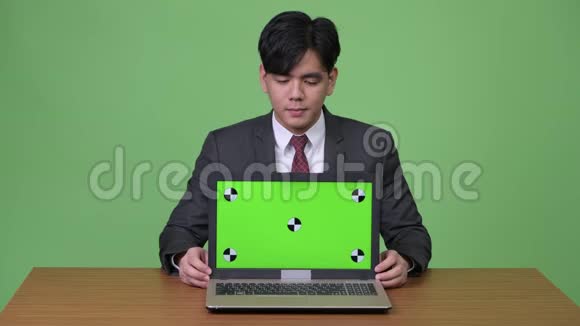 年轻英俊的亚洲商人在绿色背景下展示笔记本电脑视频的预览图
