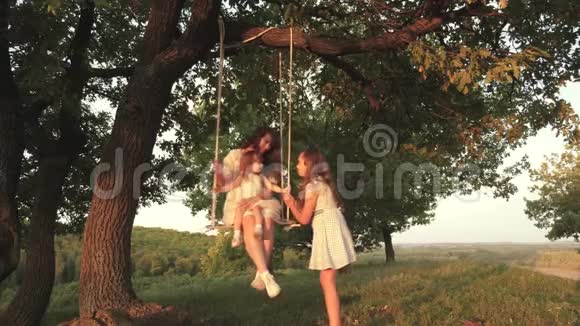 妈妈和宝宝坐在森林里的橡树树枝上的绳子秋千上在公园里的家庭乐趣在大自然中温暖的夏日母亲视频的预览图