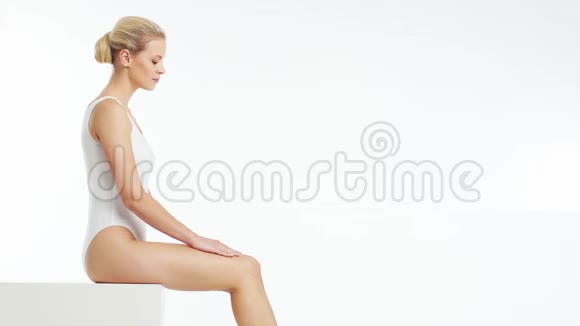年轻美丽苗条和健康的金发女孩穿着白色泳衣涂上护肤霜按摩护肤保健视频的预览图