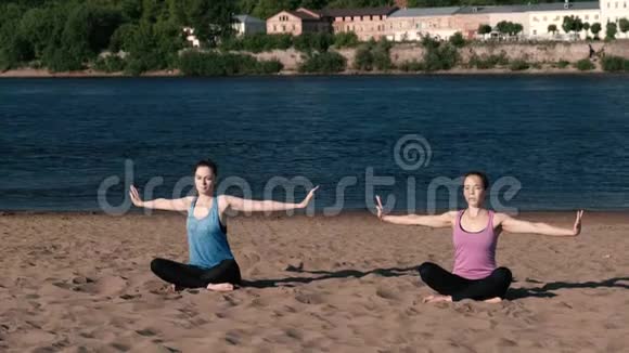 两个女人在城市河边的海滩上伸展瑜伽美丽的城市景色冥想姿势视频的预览图