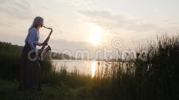 黎明时分一位年轻女子在河岸边玩萨克斯管一个穿着长裙子的漂亮女孩带着风视频的预览图
