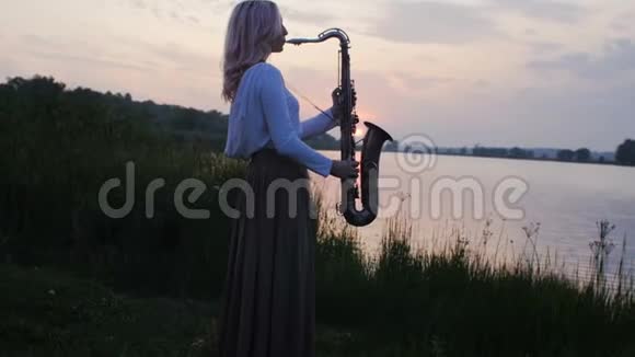 黎明时分一位年轻女子在河岸边玩萨克斯管一个穿着长裙子的漂亮女孩带着风视频的预览图