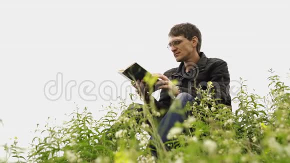 人在绿色丘陵田里看书年轻英俊的男子戴着眼镜穿着暖和的夹克在露天看书视频的预览图
