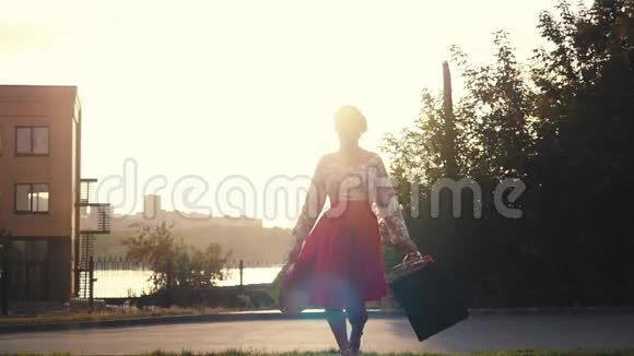 购物狂的女人穿着漂亮的连衣裙手里拿着许多购物袋在日落时分在穿过太阳视频的预览图