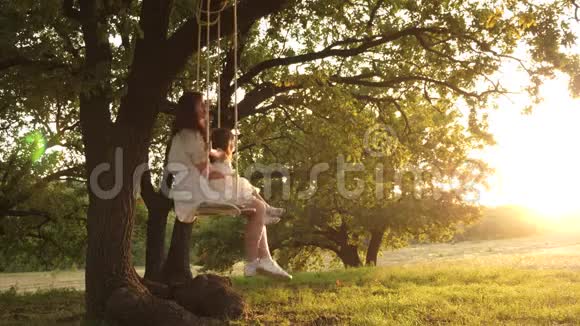 妈妈在阳光下摇动她的女儿母亲和婴儿在森林中的橡树枝上骑着一条绳秋千视频的预览图