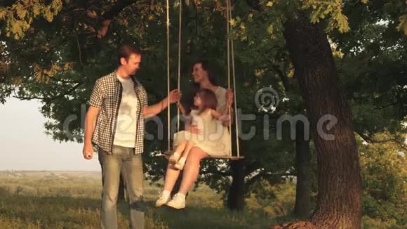 父亲摇着母亲和孩子在一根绳子上摇摆在森林里的橡树枝上妈妈摇着女儿在树下荡秋千视频的预览图