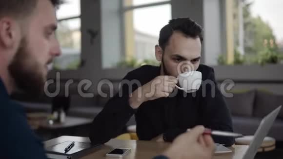 两个开朗的商人正在讨论什么一边微笑着坐在咖啡馆里拿着笔记本电脑和电话二视频的预览图