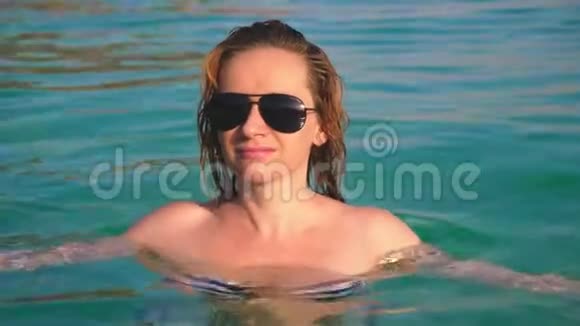 穿着泳衣和黑色太阳镜的金发女孩在蓝色海水中游泳的美丽模特视频的预览图