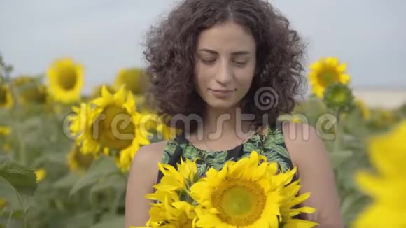 一幅美丽的卷曲嬉戏的女孩看着镜头微笑地站在向日葵田野明亮的黄色视频的预览图