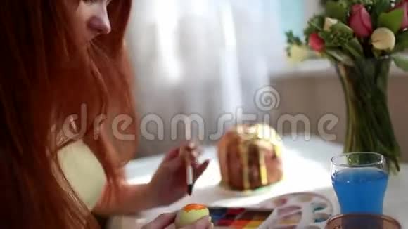红发女孩用刷子涂复活节彩蛋在桌子上复活节蛋糕鸡蛋糖果花束视频的预览图