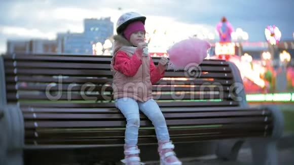 美丽的小女孩坐在游乐园的长凳上吃着滚滑轮滑冰鞋上滚来滚去的粉红色甜棉糖视频的预览图