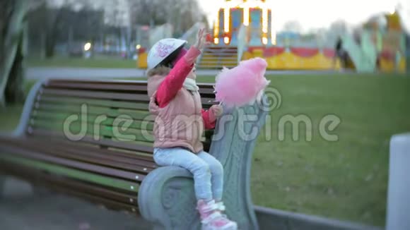 美丽的小女孩坐在游乐园的长凳上吃着滚滑轮滑冰鞋上滚来滚去的粉红色甜棉糖视频的预览图