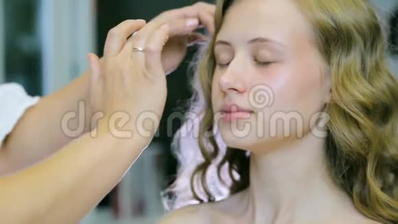 专业化妆师将奶油底涂在金色卷发和蓝眼睛模型皮肤上视频的预览图