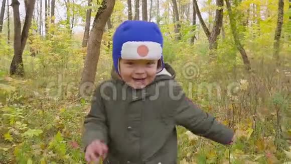 可爱的小男孩穿着绿色夹克在秋天公园的落叶中奔跑快关门视频的预览图