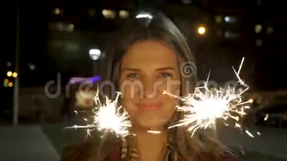 美丽微笑的年轻女子手里拿着一个闪闪发光的东西夜景年轻的微笑女孩手里拿着火花假日视频的预览图
