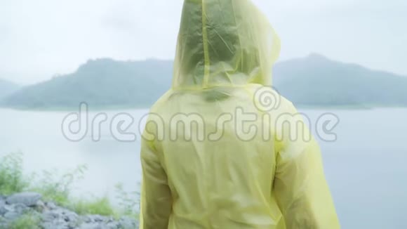 年轻的亚洲女人穿着雨衣站在湖边玩雨感到很高兴视频的预览图