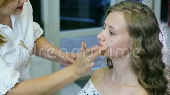专业化妆师将奶油底涂在金色卷发和蓝眼睛模型皮肤上视频的预览图