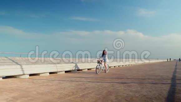 穿着蓝色连衣裙的亚洲模特骑着白色自行车在海上露台上视频的预览图