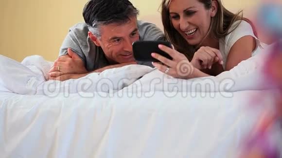 微笑的情侣躺在床上看着智能手机视频的预览图
