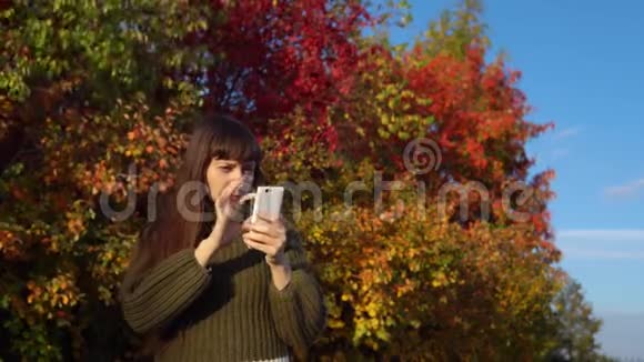 一位穿着绿色毛衣的年轻女子在秋天公园里使用智能手机视频的预览图