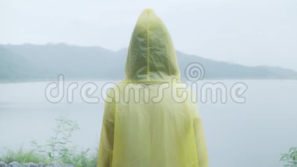 年轻的亚洲女人穿着雨衣站在湖边玩雨感到很高兴视频的预览图