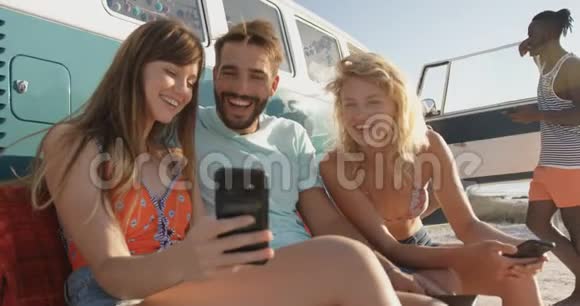 年轻的成年朋友坐在一辆露营车旁边的海滩上视频的预览图