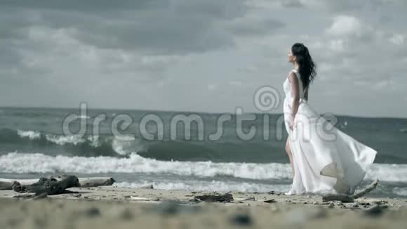 穿着白色连衣裙的漂亮女孩站在海滨女人拿着一件裙子在风中缓慢地飘动视频的预览图