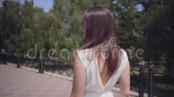 肖像迷人的年轻女孩戴着太阳镜和一件白色的夏季时装长裙走在户外休闲视频的预览图