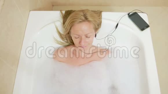 美丽的女人洗澡听耳机上的音乐手机视频的预览图