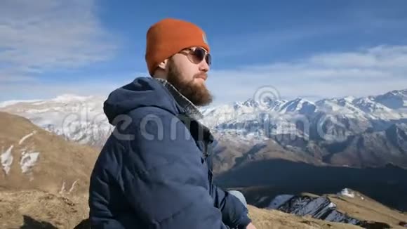 戴着太阳镜和帽子的留胡子旅行者的肖像坐在山峦背景下的岩石上视频的预览图