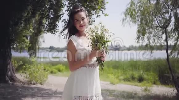 公园里站着一个年轻可爱的女孩她留着长长的黑发穿着一件白色的夏季时装长裙视频的预览图