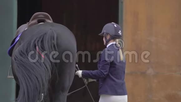 一位年轻的女骑手穿着一套黑白相间的骑师西装把她的马牵到马厩里视频的预览图